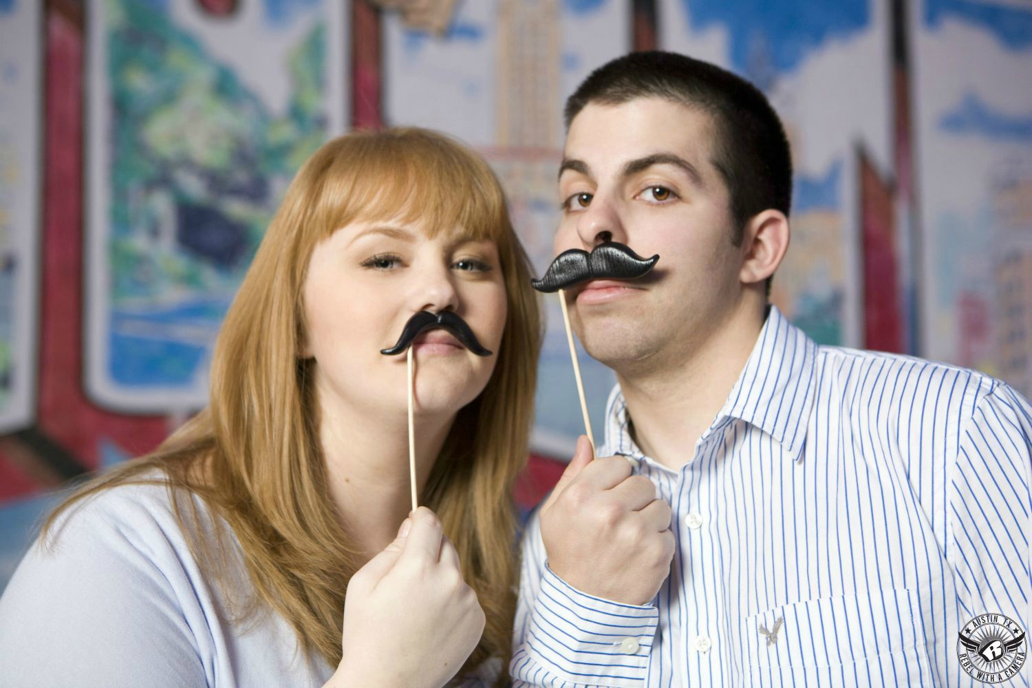 Snide, golden blond lady with a light blue blouse holds a black mustache on a stick under her nose  next to a short haired brunette guy with a white and blue striped button up dress shirt also holding a black mustache on a stick under his nose in front of the Greetings From Austin sign near SOCO in this funny engagement photograph in South Austin, Texas.